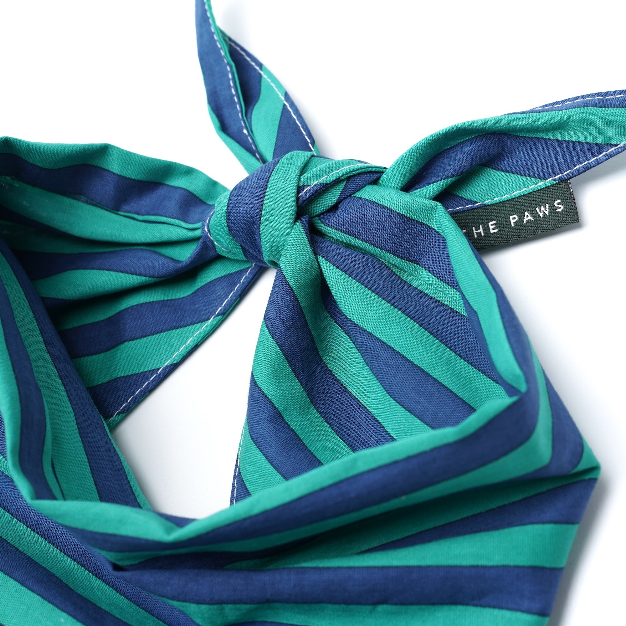 A close-up of a striped pet scarf tied in a knot, featuring alternating teal green and navy blue stripes. A black fabric brand tag reading 'THE PAWS' is visible on the side.