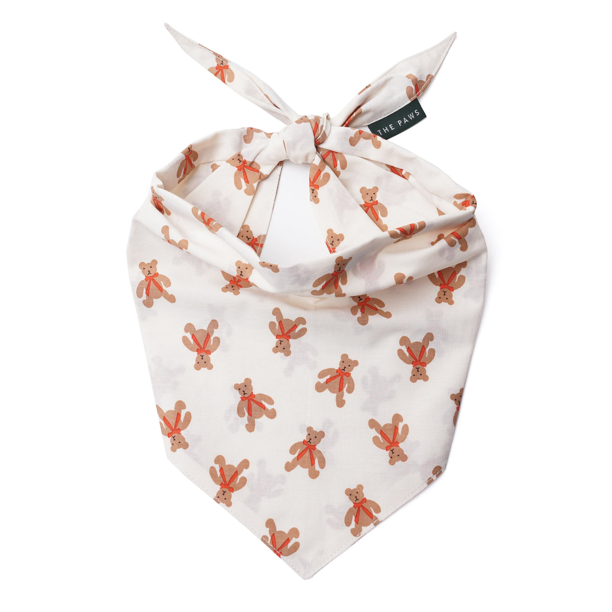 A baby bandana-style pet collar in cream with a repeating print of orange-brown teddy bears, tied at the top with a small black brand tag reading 'THE Paws'.